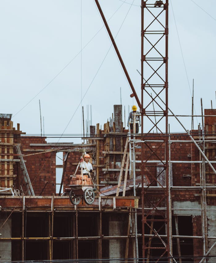 A construction worker on a scaffold at a building site with cranes and brickwork visible.